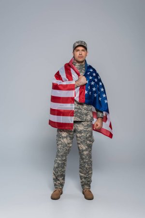in voller Länge patriotischer Armeesoldat in Tarnuniform, eingehüllt in die Flagge der Vereinigten Staaten von Amerika während des Gedenktages auf grau 