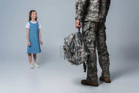 Foto de Feliz chica mirando padre en militar uniforme celebración mochila durante el regreso a casa en gris fondo - Imagen libre de derechos
