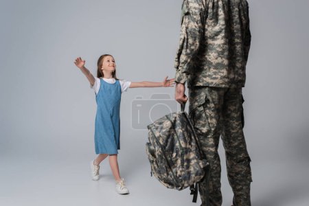 fille heureuse accueillant père en uniforme militaire avec sac à dos pendant le retour à la maison sur fond gris 