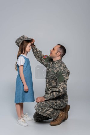 glücklicher Soldat in Armeeuniform mit Militärmütze auf dem Kopf der niedlichen Tochter auf grau 