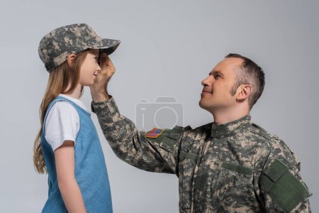 cheerful serviceman in army uniform wearing military cap on head of daughter during memorial day isolated on grey 