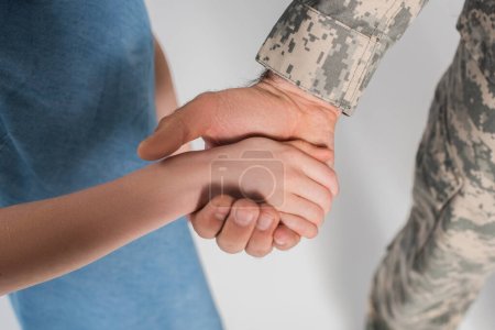 Foto de Vista recortada de niña preadolescente cogida de la mano del valiente padre en uniforme del ejército durante el día conmemorativo aislado en gris - Imagen libre de derechos