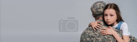 Patriotisches Mädchen umarmt Vater in Armeeuniform und weint am Gedenktag isoliert auf grauem Banner 