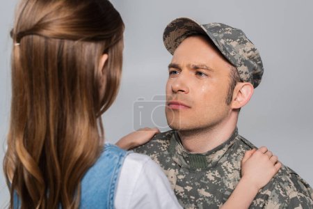 sad serviceman in military uniform crying and looking at daughter during memorial day isolated on grey 