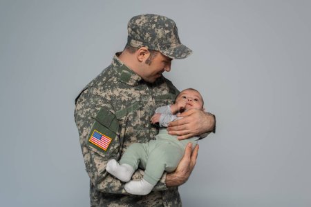 glücklicher Soldat in Uniform und Mütze, neugeborener Sohn isoliert auf grau 