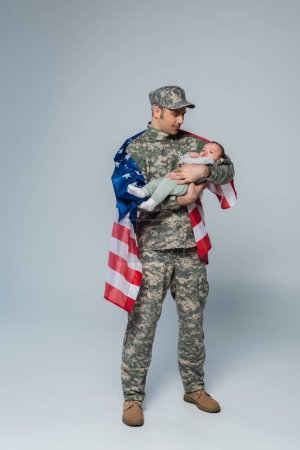 Patriotischer Soldat in Uniform und Mütze steht mit amerikanischer Flagge und hält einen kleinen Jungen in den Armen auf grau 