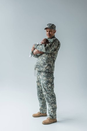 Photo for Full length of army serviceman in uniform and cap holding newborn son on grey - Royalty Free Image