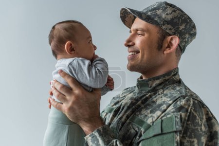 fröhlicher Armeesoldat in Uniform hält neugeborenen Sohn isoliert auf grau 