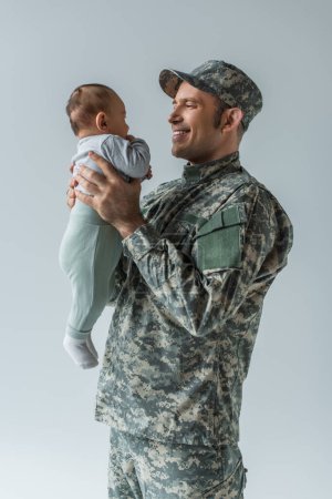 positiver Armeesoldat in Uniform mit neugeborenem Sohn im Arm isoliert auf grau 
