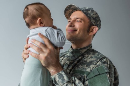 zufriedener Armeesoldat in Uniform mit neugeborenem Sohn im Arm isoliert auf grau 