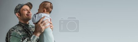 pleased soldier in military uniform holding newborn son in arms isolated on grey, banner 