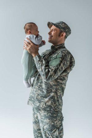 glücklicher Soldat in Uniform mit neugeborenem Sohn im Arm isoliert auf grau 
