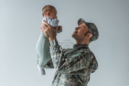 happy soldier in uniform holding baby boy in arms isolated on grey 