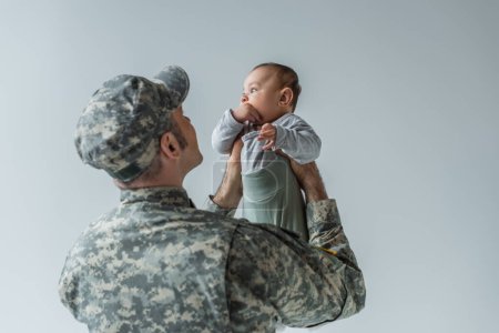 army soldier in uniform and cap holding baby boy in arms isolated on grey 