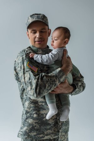 Foto de Soldado del ejército americano en uniforme militar con gorra que sostiene al niño en brazos aislados en gris - Imagen libre de derechos