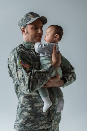 Soldado del ejército estadounidense en uniforme militar sosteniendo a su hijo en armas aislado en gris