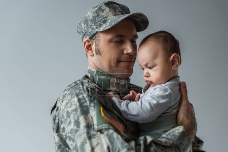 Soldado americano en uniforme militar sosteniendo en armas hijo recién nacido aislado en gris