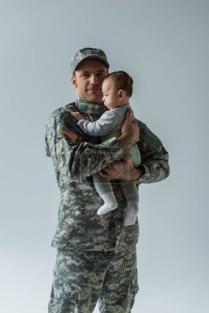 pleased American serviceman in military uniform holding in arms newborn son isolated on grey