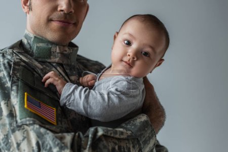 Hombre de servicio americano en uniforme sosteniendo en brazos niño recién nacido aislado en gris