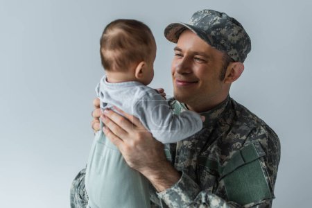 fröhlicher Soldat in Uniform hält neugeborenen Jungen in den Armen, isoliert auf grau 
