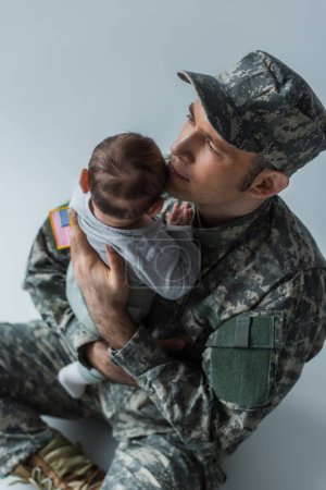 vista de ángulo alto del soldado de los E.E.U.U. en uniforme que sostiene en brazos al muchacho recién nacido mientras que está sentado en gris 