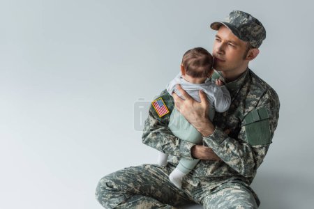 proud father in military uniform holding in arms newborn son while sitting on grey background 