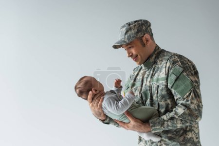 glücklicher US-Soldat in Uniform schaut neugeborenen Jungen an und lächelt isoliert auf grau 