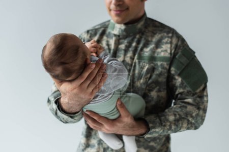 Foto de Vista recortada del orgulloso padre en uniforme militar sosteniendo en armas hijo recién nacido aislado en gris - Imagen libre de derechos
