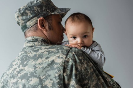 Vater in Militäruniform und Mütze umarmt neugeborenen Jungen isoliert auf grau 