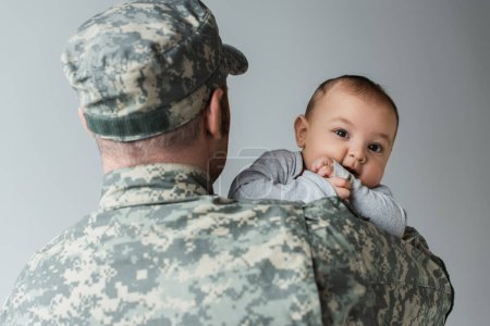 serviceman in military uniform and cap hugging newborn son isolated on grey 