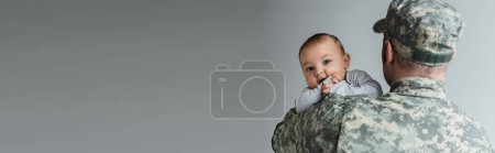 serviceman in military uniform and cap hugging newborn son isolated on grey, banner 