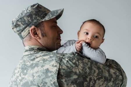 fröhlicher Vater in Militäruniform und Mütze umarmt neugeborenen Sohn isoliert auf grau 