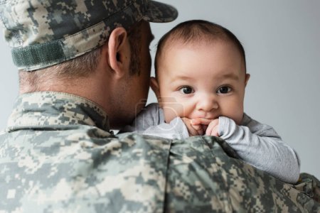 Mann in Armeeuniform und Mütze umarmt neugeborenen Sohn isoliert auf grau 