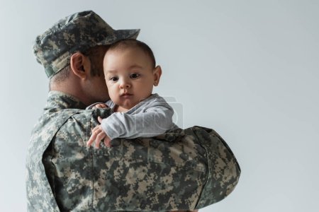 father in military uniform and cap hugging infant son isolated on grey 