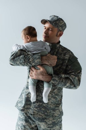 stolzer Soldat in Militäruniform und Mütze umarmt kleinen Sohn isoliert auf grau 