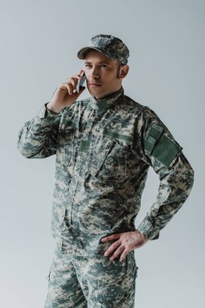 Soldat in Mütze und Armeeuniform unterhält sich auf Smartphone isoliert auf grau 