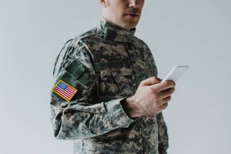 Foto de Vista recortada del militar estadounidense utilizando un teléfono inteligente aislado en gris - Imagen libre de derechos