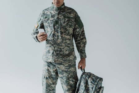 Ausgeschnittene Ansicht eines amerikanischen Soldaten, der sein Smartphone benutzt, während er seinen Rucksack isoliert auf grau hält 