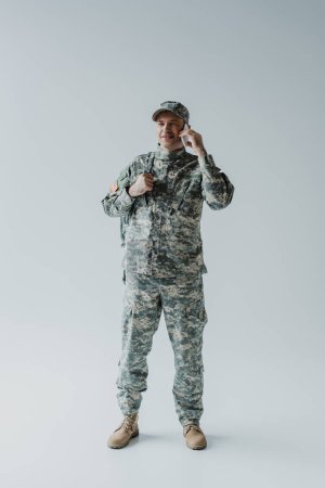 Téléchargez les photos : Pleine longueur de militaire heureux en casquette et uniforme parler sur smartphone sur gris - en image libre de droit