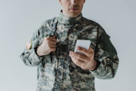 vista recortada del militar en uniforme del ejército utilizando el teléfono móvil aislado en gris 
