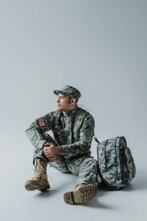 Soldat américain en uniforme de l'armée avec drapeau national assis près du sac à dos sur gris 
