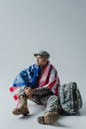 American soldier in army uniform and national flag of United States sitting near backpack on grey 