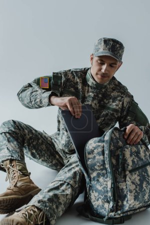 Amerikanischer Soldat in Armeeuniform und Nationalflagge der Vereinigten Staaten nimmt Laptop aus Rucksack auf grau 