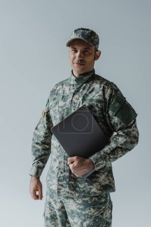 good looking soldier in army uniform and cap standing with laptop isolated on grey 