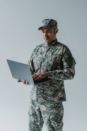 good looking soldier in army uniform and cap using laptop isolated on grey 