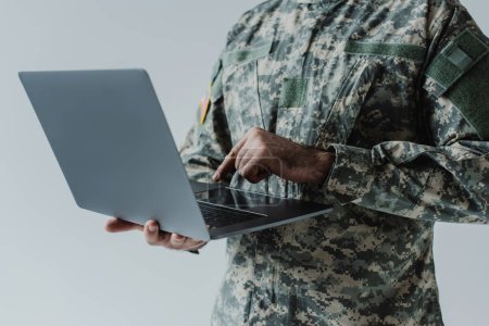 Ausgeschnittene Ansicht eines Soldaten in Armeeuniform mit Laptop isoliert auf grau 