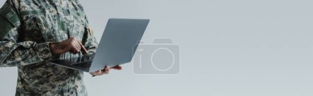 Ausgeschnittene Ansicht eines Soldaten in Armeeuniform mit Laptop isoliert auf grau, Banner 