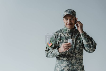 soldier in uniform and cap holding earphone case and smiling isolated on grey 