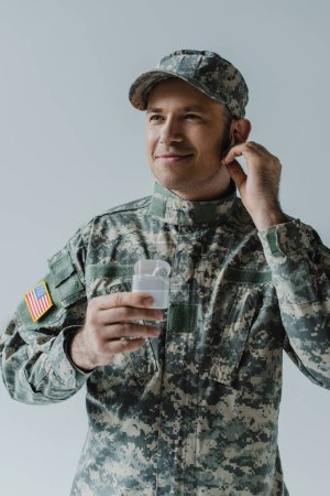 soldat joyeux en uniforme et casquette tenant le boîtier de l'écouteur et souriant isolé sur gris 