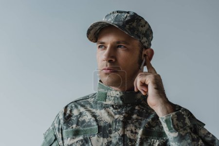 soldier in uniform and cap adjusting wireless earphone isolated on grey 